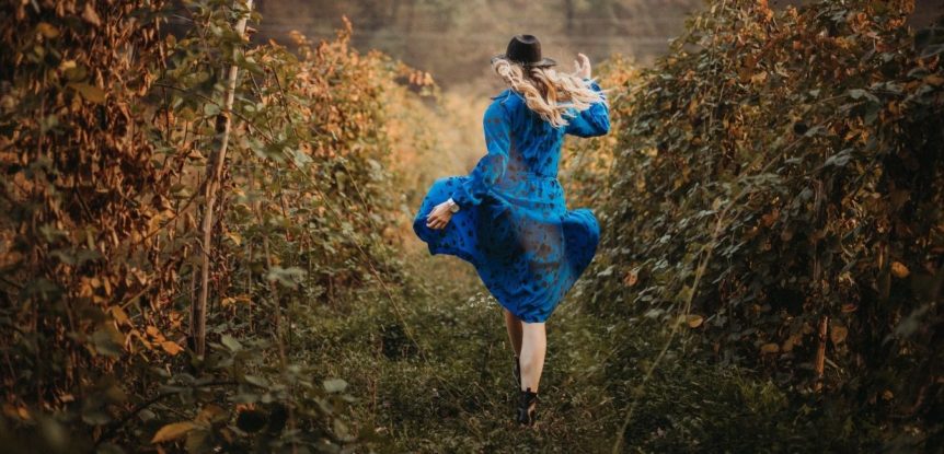 She was having so much fun, her family was worried. Woman in a blue dress and black hat skipping through a field.